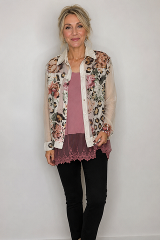 A woman modeling a sheer cream button-up shirt with a mixed floral and leopard print pattern, worn open over a dusty rose top and black pants, against a plain white wall.